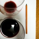 A pair of wine glasses viewed from above. One glass has less red wine than the other. A charcoal pencil rests to the right of the glassware.